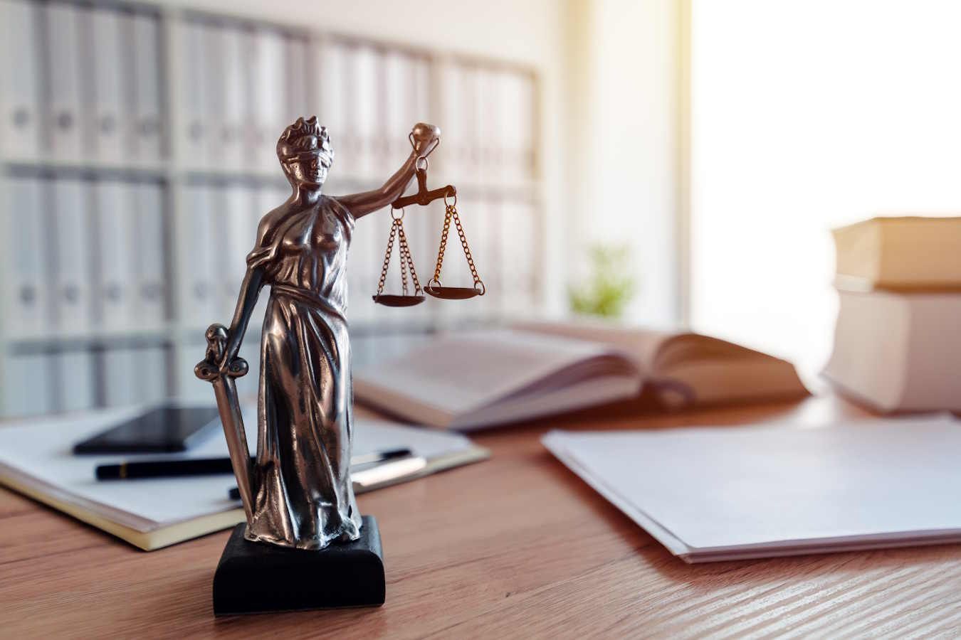 Eine Justitia Statue auf einem Holztisch. Im Hintergrund liegen Bücher und Dokumente. Als Anwalt für Verwaltungsrecht im Meppen vertreten wir Sie bei rechtlichen Angelegenheiten.