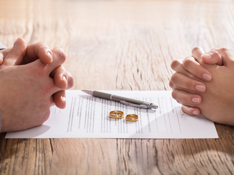 Rechts und links je zwei gefaltete H&auml;nde, zwischen ihnen liegt ein Dokument, auf dem ein Kugelschreiber und zwei Eheringe liegen. Ein Termin bei einem Scheidungsanwalt sorgt f&uuml;r eine faire Scheidung. 